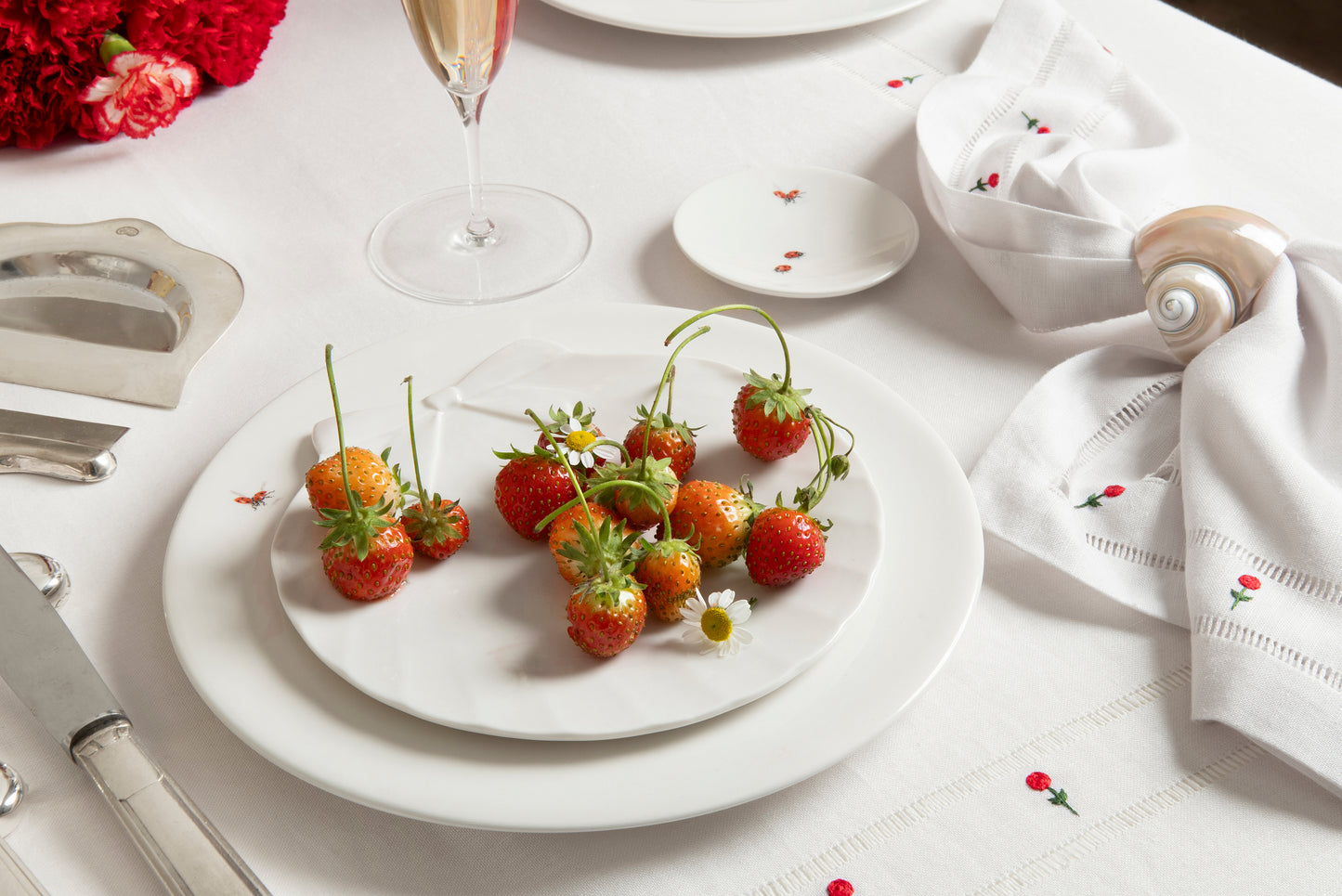 White table setting with strawberries on a plate, red flowers, and silver cutlery.